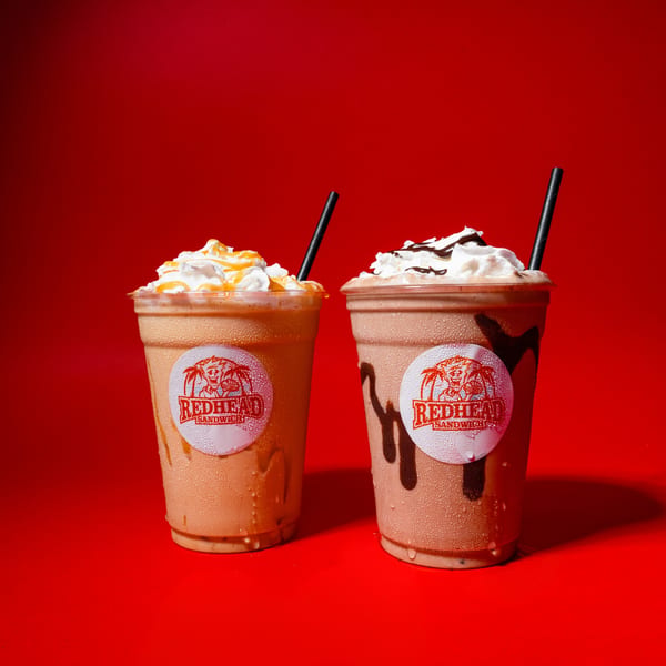 Two Redhead Sandwich milkshakes, one caramel and one chocolate, with whipped cream and straws on a red background.