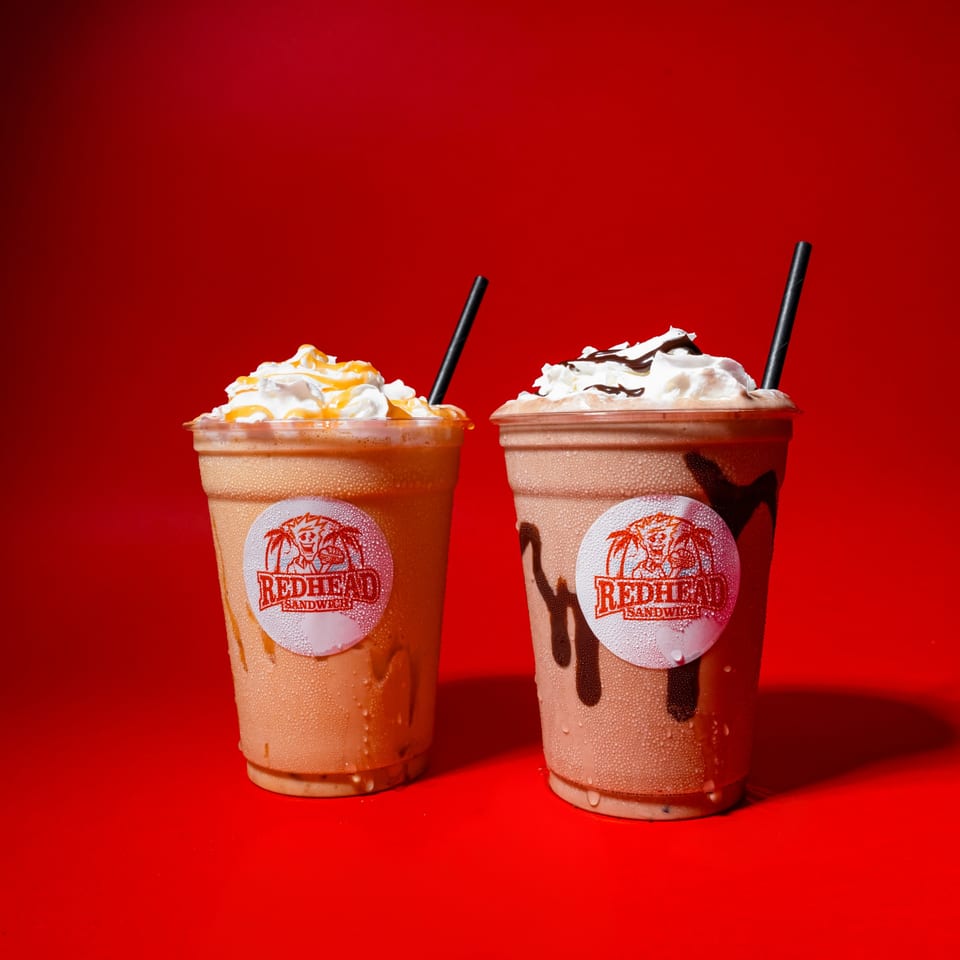 Two Redhead Sandwich milkshakes, one caramel and one chocolate, with whipped cream and straws on a red background.