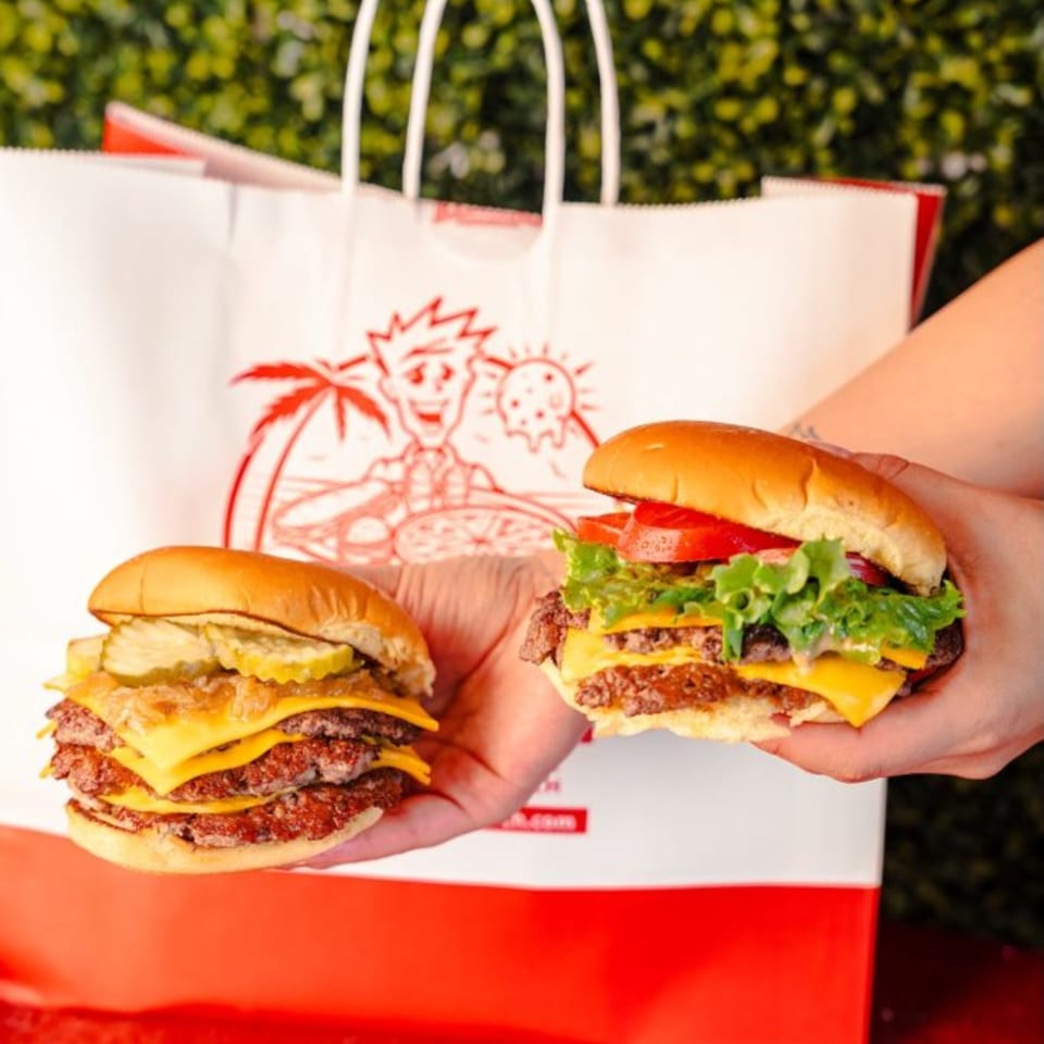 Two hands hold two RedHead Burger double cheeseburgers, one with pickles and onions, the other with lettuce and tomato.